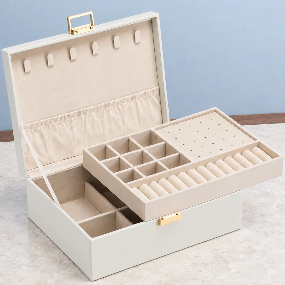 VLANDO — coffret beige à double couche de VLANDO, mise en situation dans un décor de pierre perle claire et de bleu ardoise pâle — Vue éclatée du plateau amovible au-dessus du coffret ; le plateau repose dans la boîte en usage.